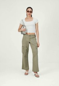 Γυναίκα φορώντας λευκό crop top, ελαιόχρωμες cargo παντελόνες, λευκές γόβες με τακούνι, γυαλιά ηλίου, και κρατώντας μια μικρή γκρίζα τσάντα χεριού.