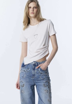 Młoda kobieta o długich blond włosach, ubrana w biały krótki top z krótkim rękawem oraz niebieskie jeansy z wysokim stanem ozdobione metalicznymi ćwiekami i wzorami gwiazd.