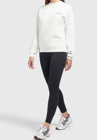 Personne portant un sweat-shirt blanc avec un petit texte, des leggings noirs et des chaussures de course blanches avec des accents noirs et orange, marchant vers l'avant.