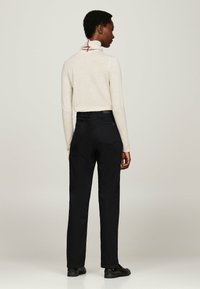 Bež turtleneck-sviiter punase ja valge detailiga seljaosas, koos mustade sirgetest pükstest, vaadates tagant.