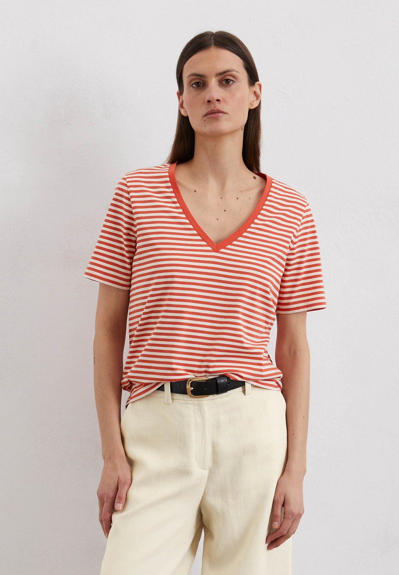 Gestreiftes V-Ausschnitt-T-Shirt mit orange- und weißen Streifen, kurzen Ärmeln und einer lockeren Passform. Kombiniert mit cremefarbenen Hosen und einem schwarzen Gürtel.
