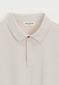 Polo beige en tissu lisse avec un col classique, une patte de boutonnage à deux boutons et un bouton d'accent noir. Étiquette de la marque visible.