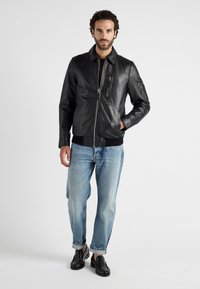 Schwarze Lederbomberjacke mit Reißverschluss, geripptem Saum und zwei Reißverschlusstaschen. Darunter werden hellblaue Denim-Jeans und schwarze Slipper getragen.