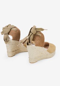 Sandalias de cuña con parte superior de gamuza color marrón, tacón de paja trenzada y cintas caqui en el tobillo. Diseño de espalda abierta y puntera redondeada.