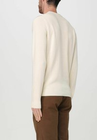 Maglione lavorato a maglia color crema con scollatura rotonda e polsini a coste, indossato con pantaloni marroni, che mostrano una vista posteriore semplice.