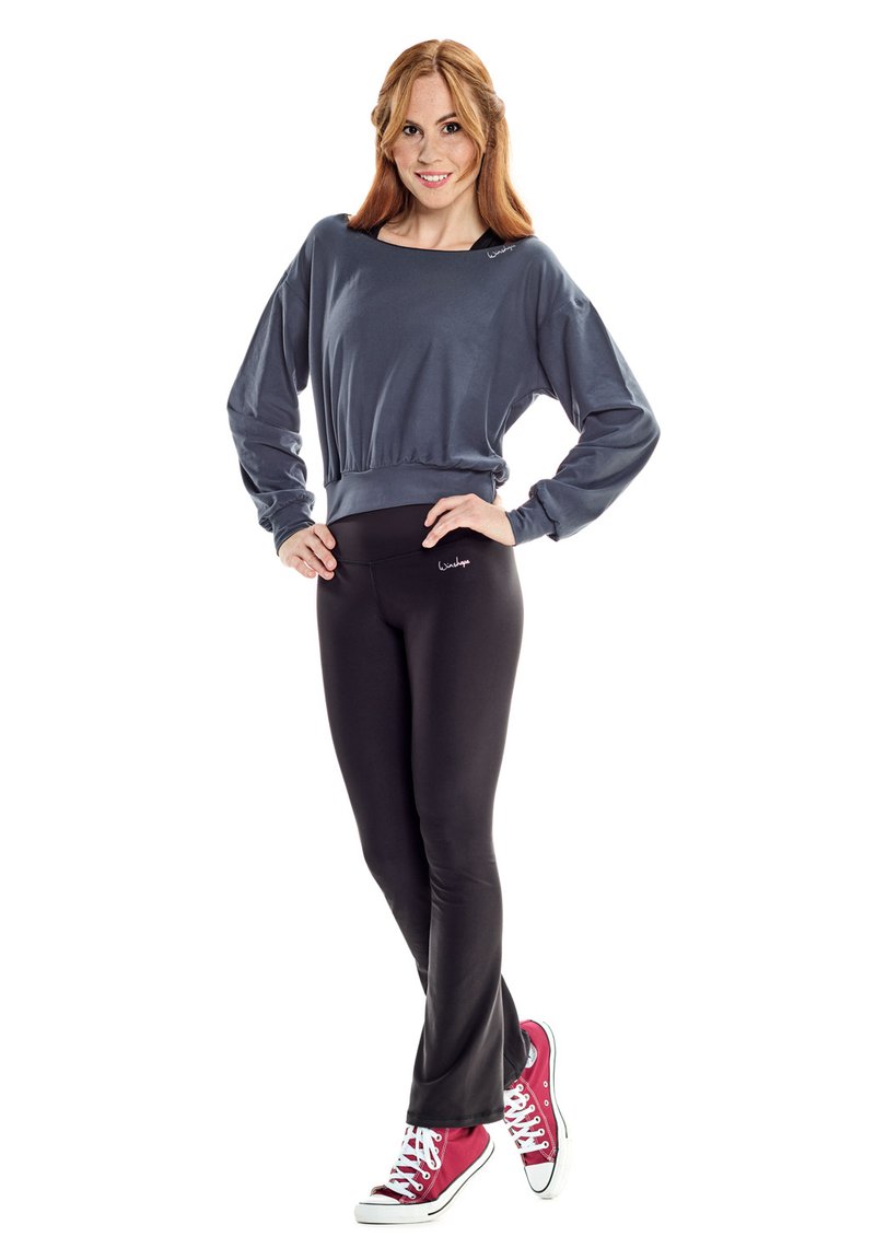 Grauer, kurz geschnittener Sweatshirt mit langen Ärmeln, schwarze, enganliegende Leggings und rote Sneakers mit weißen Schnürsenkeln. Die Leggings haben ein Logo.