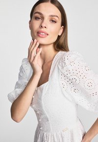 Biała sukienka z haftem, z bufiastymi rękawami, z kwadratowym dekoltem, z ozdobnymi guzikami i złotą metalową akcentacją. Gładki, fakturowany materiał.