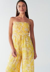 Jumpsuit sans bretelles jaune avec une bodice froncé, une taille élastique et un motif de feuilles. Tissu léger avec une silhouette fluide.
