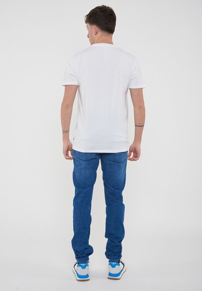 T-shirt bianca a maniche corte, abbinata a jeans in denim blu. Sneakers con accenti blu e grigi su uno sfondo neutro.