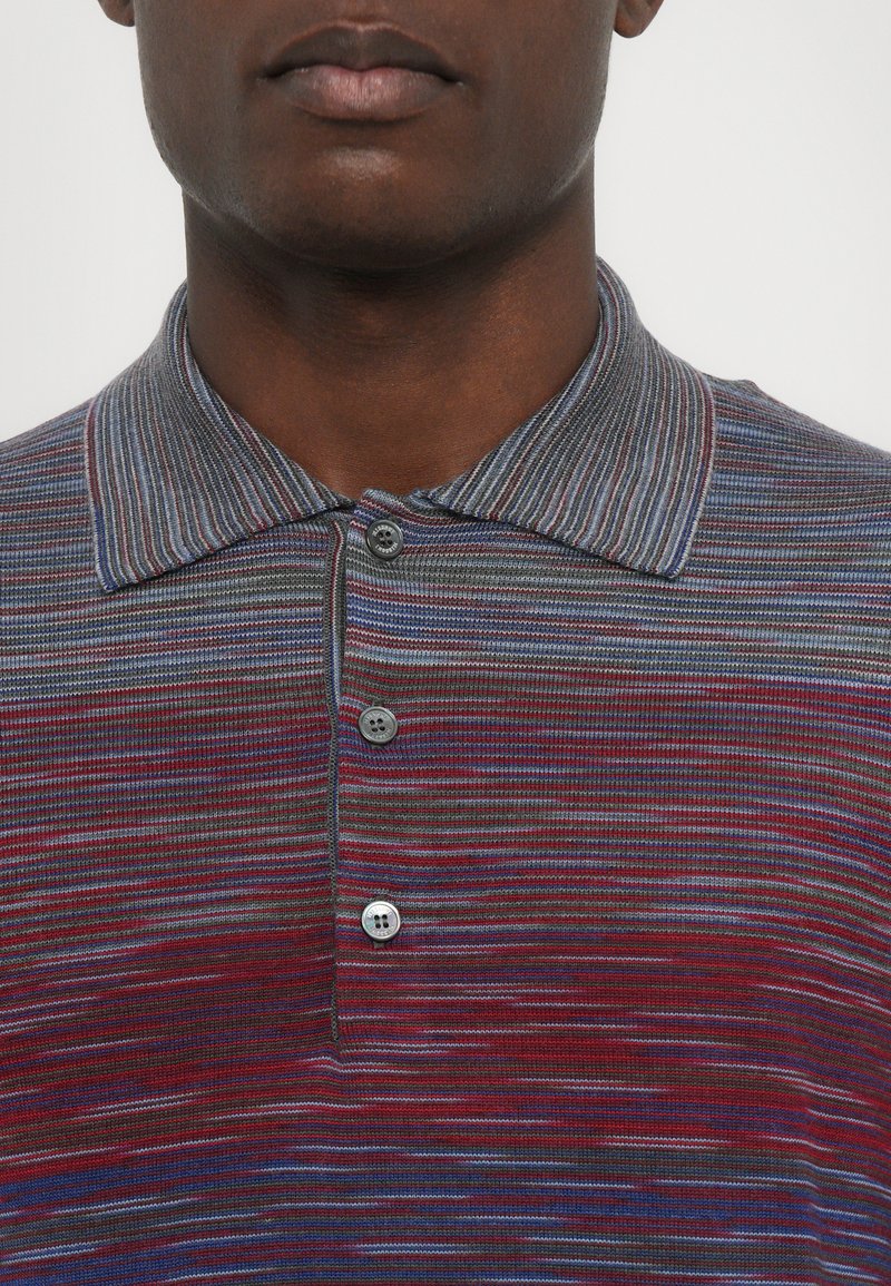 Camisa polo a rayas con cuello acanalado, botones en el frente y una paleta de colores en rojo oscuro, azul y gris. Textura suave.