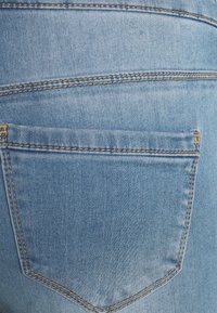 Jeans de mezclilla en azul claro, con un bolsillo trasero con costuras amarillas y una textura suave. Diseño clásico de cinco bolsillos.