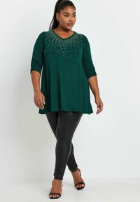 Vihreä tunika, jossa on V-pääntie ja koristellut niitit olkapäillä, yhdistetty mustiin legginsseihin ja nauhakorkkareihin. Siro kangas, rento istuvuus.