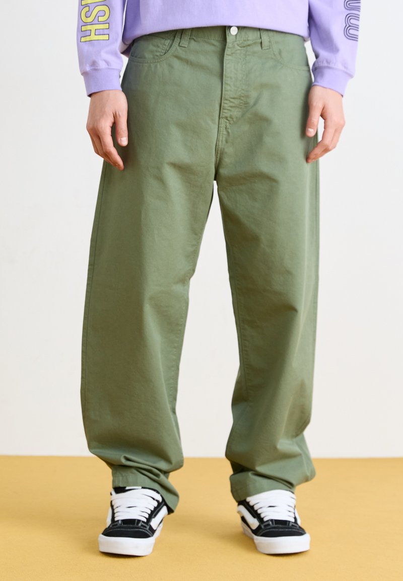 Pantalones de algodón verdes holgados con grandes bolsillos, combinados con zapatillas negras y blancas, llevados sobre una camisa de manga larga lavanda.