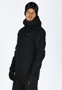 Schwarze Winterjacke mit Kapuze und mehreren Reißverschlusstaschen. Glatter, strapazierfähiger Stoff und enganliegende Bündchen. Getragen mit schwarzen Handschuhen.