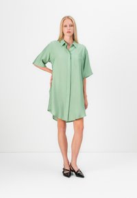 Rochie de cămașă cu mânecă scurtă, de culoare verde deschis, cu deschidere prin butoane și buzunar. Țesătura pare netedă, cu o croială ușor lejeră și margine curbată.