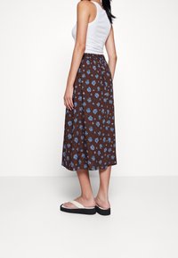 Falda midi marrón con estampado de flores azules, cintura elástica y corte holgado. Se lleva con sandalias planas sobre un fondo liso.