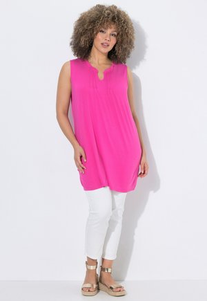 Femme aux cheveux bouclés portant une tunique sans manches rose vif, un pantalon blanc slim et des sandales compensées dorées, debout devant un fond blanc.