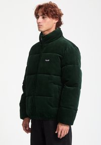 Green corduroy puffer jacket with high collar, zip closure, and visible stitching. Features a logo on the chest and ribbed cuffs.