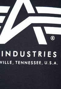 Close-up van een donkere stof met vetgedrukte witte letters die "INDUSTRIES" en "VILLE, TENNESSEE, U.S.A." spellen, met daarboven een gedeeltelijk geometrisch logo.