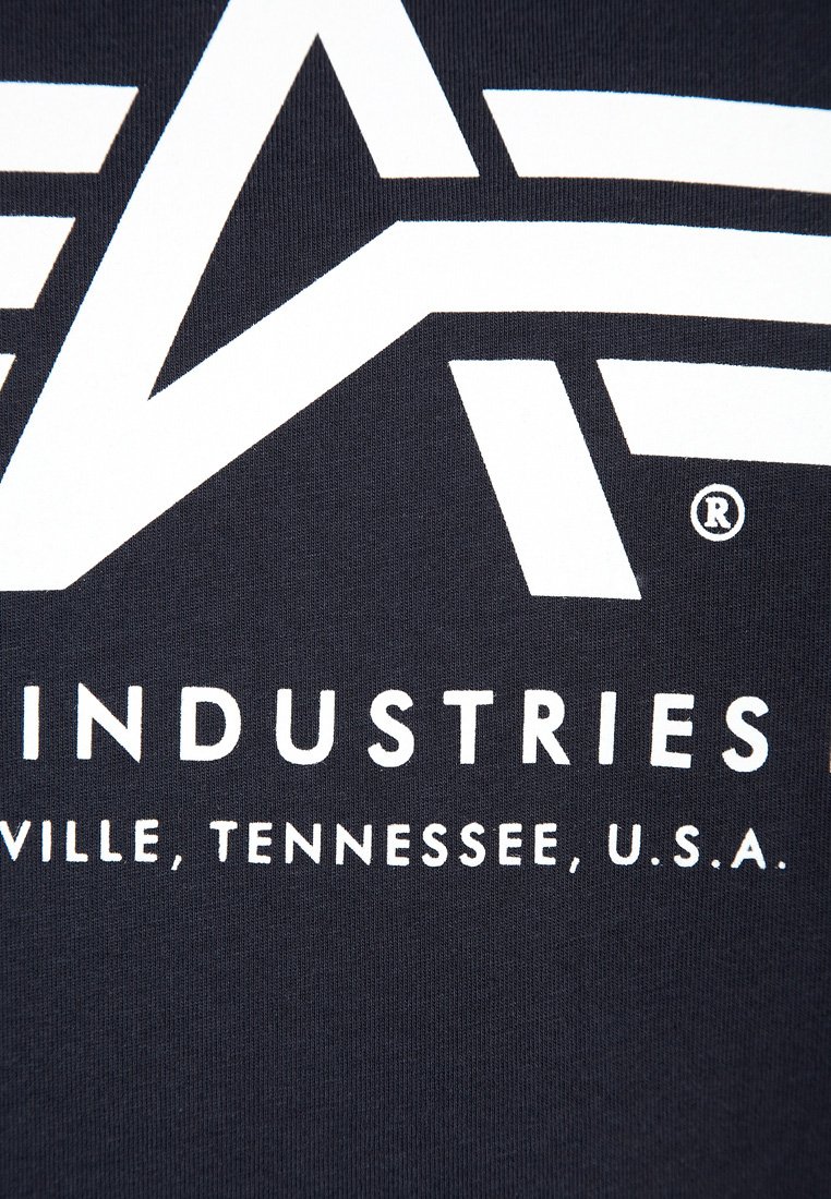 Zbliżenie na ciemny materiał z wyraźnymi białymi literami, na których widnieje napis "INDUSTRIES" oraz "VILLE, TENNESSEE, U.S.A." z częściowym geometrycznym logo powyżej.