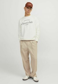 Felpa bianca con stampa nera "Ocean club", pantaloni beige plissettati e berretto marrone. Sneakers bianche con accenti grigi e neri.