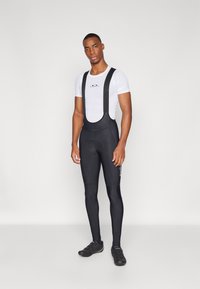 Bib shorts da ciclismo neri con bretelle sopra una maglietta in mesh a maniche corte bianca. Presentano un sedile imbottito e un marchio sul lato.