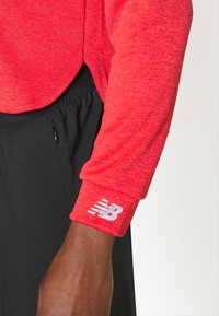 Maglia atletica rossa a maniche lunghe con un tessuto texturizzato. I polsini presentano un logo bianco di New Balance. Abbinata a pantaloni corti neri con coulisse.