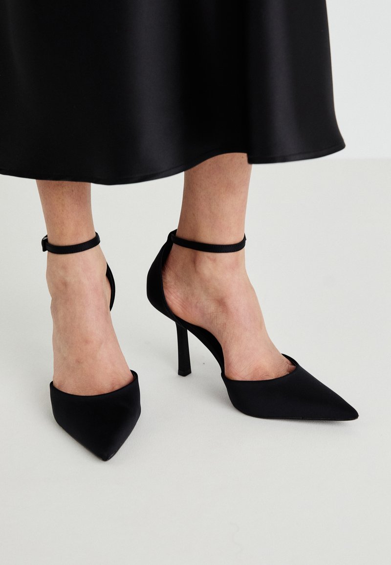 Schwarze spitze High Heels aus glattem Stoff mit einem Stiletto-Absatz und einem schmalen Knöchelriemen, mit einem offenen Seitendesign.