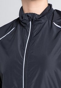 Chaqueta negra con cremallera, fabricada en un material ligero, que presenta un cuello alto y rayas blancas de acento a lo largo de los hombros y la cremallera.