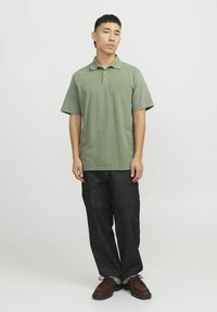 Polo verde con una trama morbida, maniche corte e colletto, abbinato a pantaloni in denim scuri e scarpe marroni. Emblema semplice sul petto.