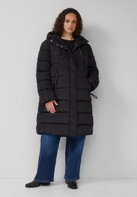 Schwarze Pufferjacke mit Kapuze, horizontaler Nahtstiche, Reißverschlusstaschen und verstellbaren Zugbändern in der Taille, kombiniert mit blauen Schlagjeans.