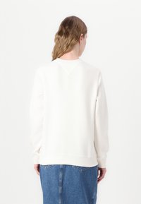 Hvit genser med lange ermer, rund hals og ribbestrikket kant. Glatt, myk tekstur. Brukt sammen med et denimskjørt, sett fra baksiden.