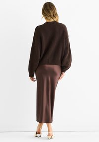 Brown knit oversized sweater with ribbed hem, paired with a fitted brown satin midi skirt and cream-colored heeled sandals.