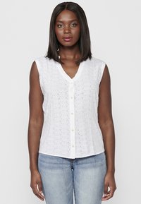 Blouse sans manches blanche en tissu œillets, décollet�é en V et devant à boutons. Présente une texture subtile et des motifs floraux découpés. Portée avec un jean bleu.