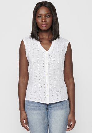 White sleeveless blouse with eyelet fabric, v-neck, and button front. Features subtle texture and floral cut-out patterns. Worn with blue jeans.
