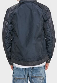 Man som bär en svart vadderad läderjacka med sömdetaljer på ärmarna och axlarna, tillsammans med blå denimjeans.