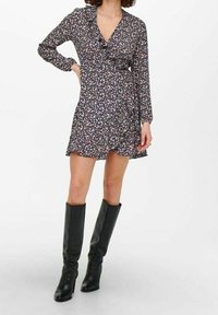 Žena v dlouhém černém květinovém zavinovacím šatech s volány, v kombinaci s černými koženými vysokými botami na podpatku, stojící před bílým pozadím.