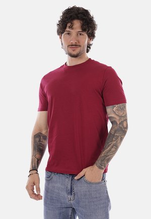 Uomo con capelli ricci e tatuaggi sulle maniche, che indossa una maglietta rossa e jeans blu, in piedi con una mano in tasca su sfondo bianco.