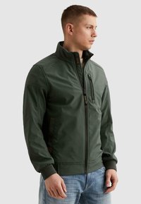 Man in een groen jack met ritssluiting en hoge kraag, met een ritssluiting op de borstzak, draagt lichtblauwe spijkerbroek, kijkt naar rechts tegen een effen achtergrond.