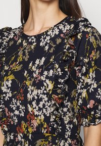 Robe florale navy avec des fleurs blanches, jaunes et roses, ornée d'un volant le long des manches bouffantes et de la poitrine.