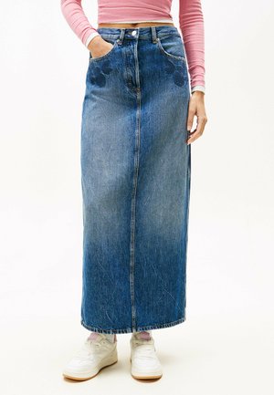 Falda maxi de mezclilla en azul medio con corte recto, textura descolorida, bolsillos y bordado floral en el frente. Combinada con zapatillas blancas.
