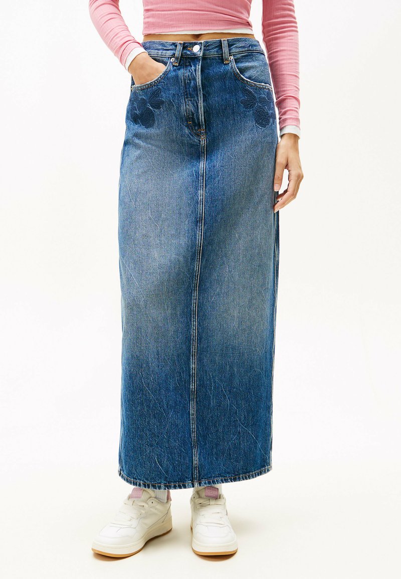Gonna maxi in denim blu medio con taglio dritto, texture sbiadita, tasche e ricami floreali sul davanti. Abbinata a sneakers bianche.