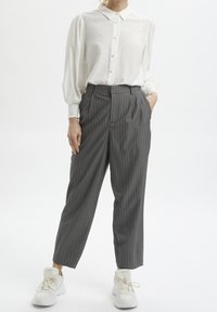 Biała koszula z guzikami i bufiastymi rękawami wpuszczona w szare spodnie w paski. Model nosi białe trampki. Gładki materiał i dopasowany krój.