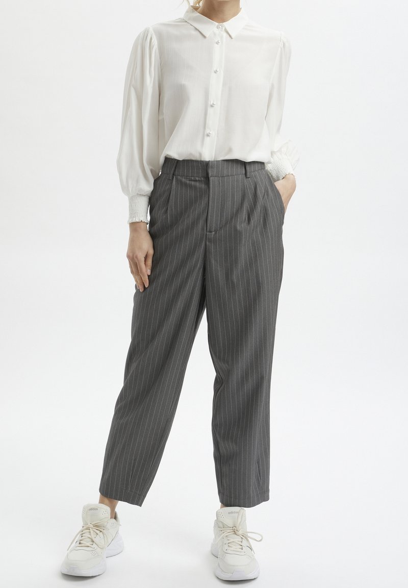 Biała koszula z guzikami i bufiastymi rękawami wpuszczona w szare spodnie w paski. Model nosi białe trampki. Gładki materiał i dopasowany krój.