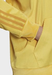 Gul manchesteroveralljacka med ribbad textur, design med tre ränder på ärmarna och en framficka. Mjukt tyg med ett mysigt utseende.