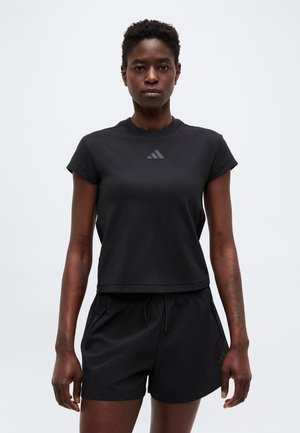 Femme debout, face à l'avant, portant un t-shirt de sport noir à manches courtes et un short noir à cordon, sur un fond uni.