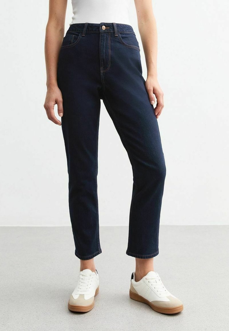 Jean en denim bleu foncé à taille haute, coupe droite, avec des détails de couture subtils et des poches avant. Associé à des baskets blanches.