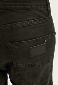 Črne jeans hlače iz denima, ki imajo žep z pravokotnim usnjenim označevalcem. Tkanina ima gladko teksturo z vidnimi šivi.