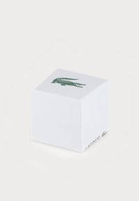 Baltas popierinis dėžutė su tekstūruota paviršiumi, ant viršaus esantis žalias krokodilo logotipas ir šone užrašas "LACOSTE".