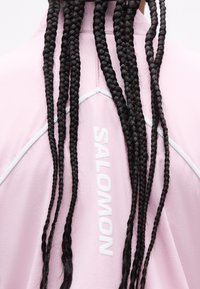 Μακριές μαύρες κοτσίδες κρέμονται πάνω από ένα ανοιχτό ροζ ένδυμα με λευκό κείμενο "SALOMON" και λευκές ραφές στο πίσω μέρος.